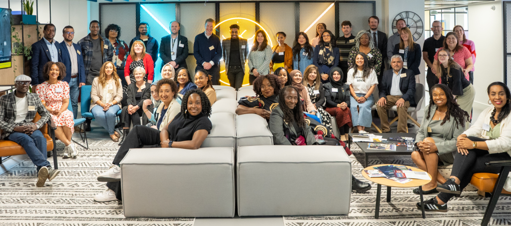 a group of 42 people are standing or sitting on white sofas looking to camera smiling
